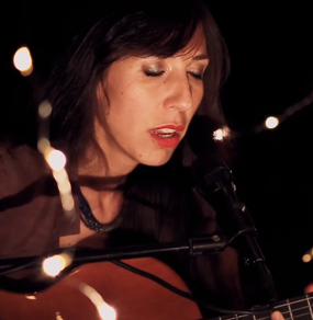 Anna Vignozzi canta un brano live con chitarra e occhi chiusi, in esterni notturni, con lucine d'atmosfera
