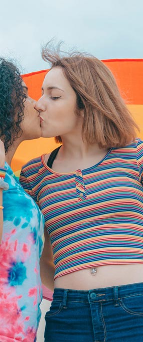 Due ragazze con magliette colorate, una creola e l'altra bianca, si baciano sullo sfondo della bandiera Lgbt