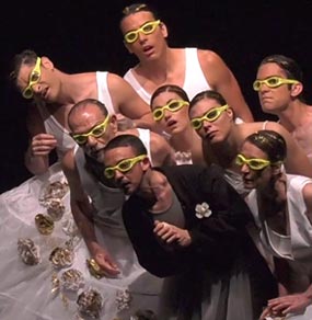 Un gruppo di attori con abiti bianchi, tranne quello in primo piano con giacca nera, e occhiali da piscina gialli